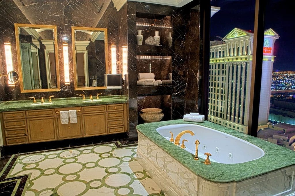 Luxurious bathroom with marble surfaces, a green-topped vanity, large bathtub, mirrors, and a night view of city buildings through floor-to-ceiling windows.