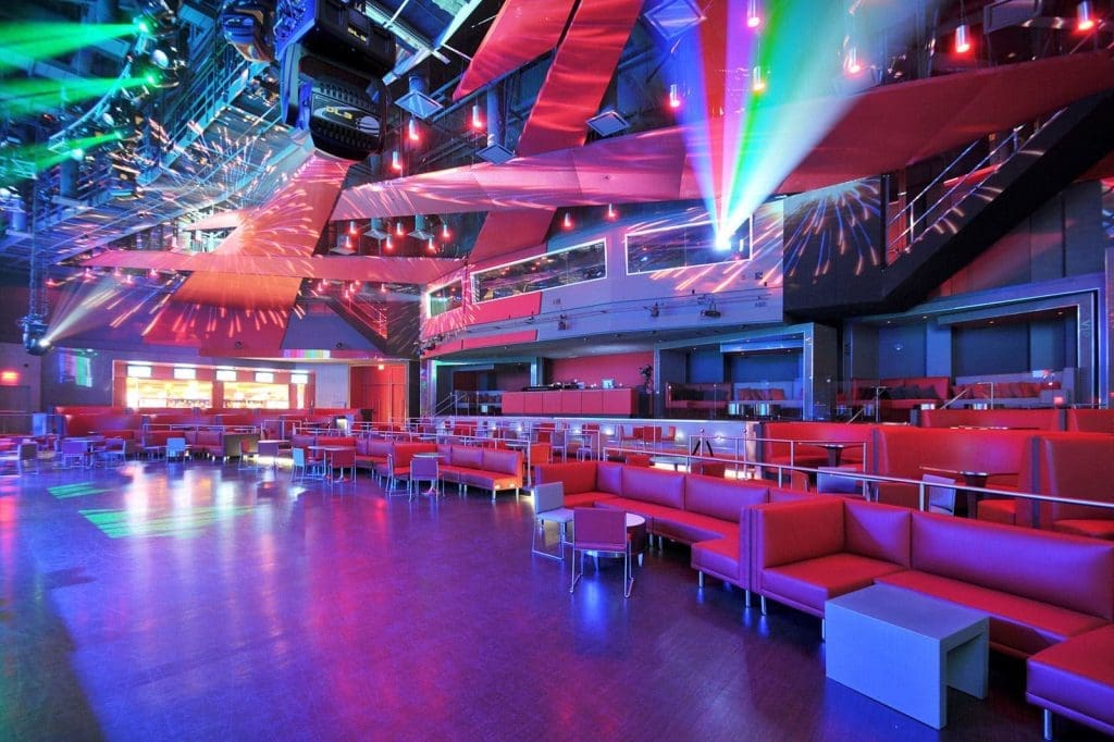Large modern nightclub interior with red seating, colored lights, reflective ceiling, and spacious dance floor.