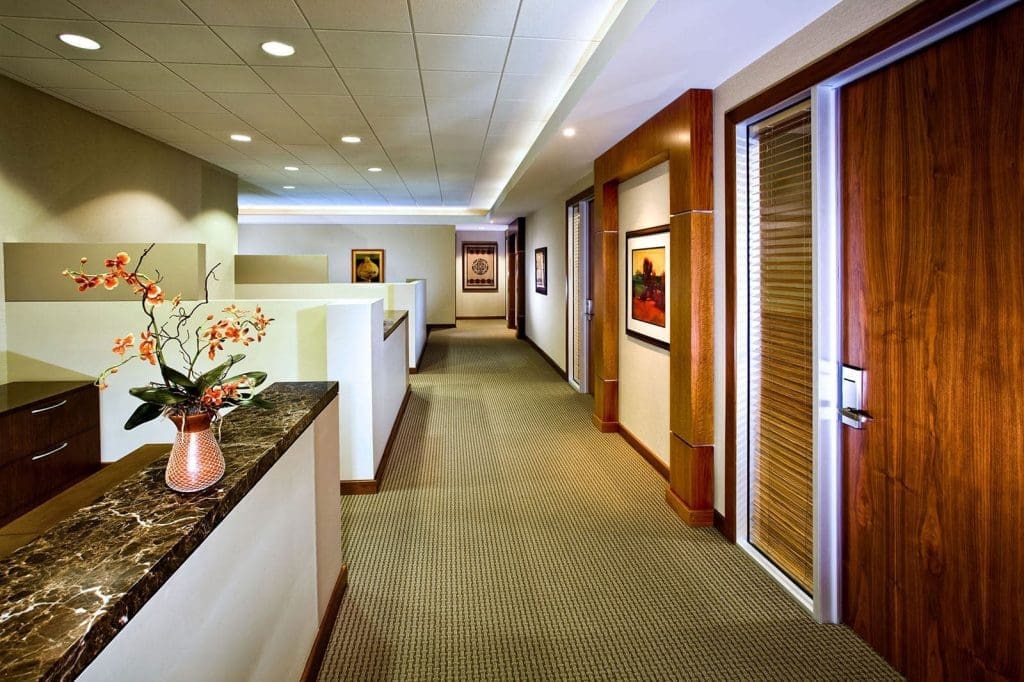 Modern office hallway with beige carpet, wooden doors, framed art on walls, and a marble counter with a vase of orange flowers in the foreground.