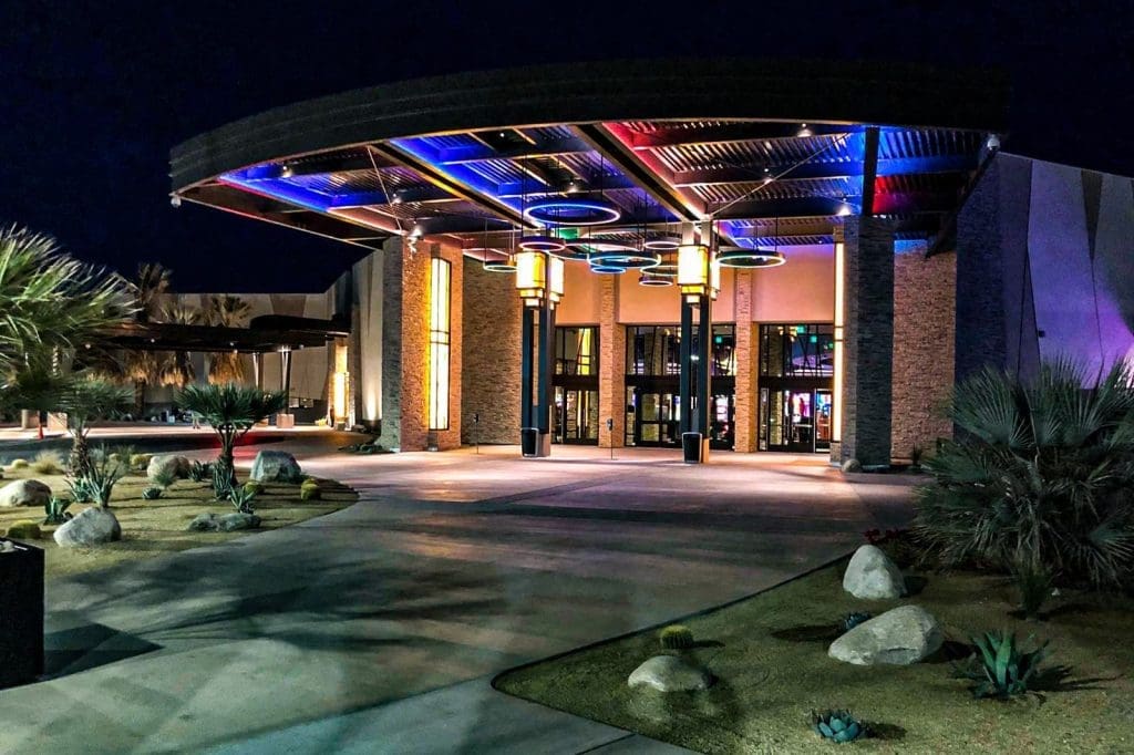 The entrance to a modern building at night, featuring colorful overhead lights, large glass doors, and landscaped desert plants.