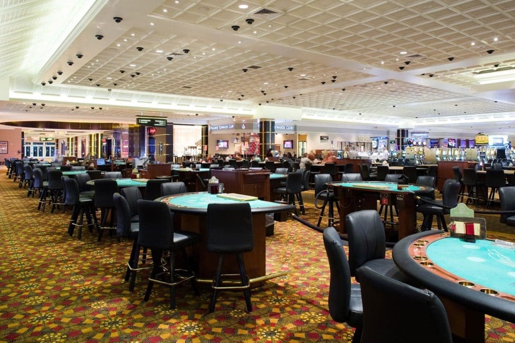 A spacious casino floor with empty gaming tables and chairs, patterned carpet, and bright overhead lighting. Slot machines and signs are visible in the background.