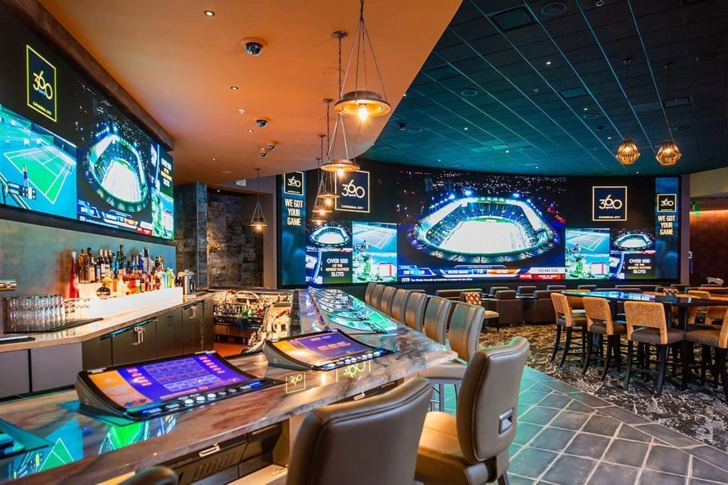 Modern sports bar with large screens displaying sporting events, a long bar with stools, and a row of betting kiosks on the counter.