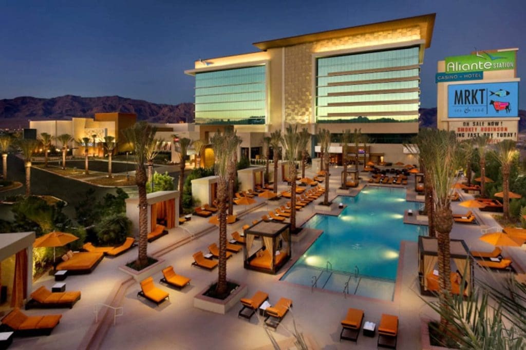 Outdoor view of Aliante Casino and Hotel at dusk, featuring a modern building, illuminated pool area, orange lounge chairs, palm trees, and mountains in the background.