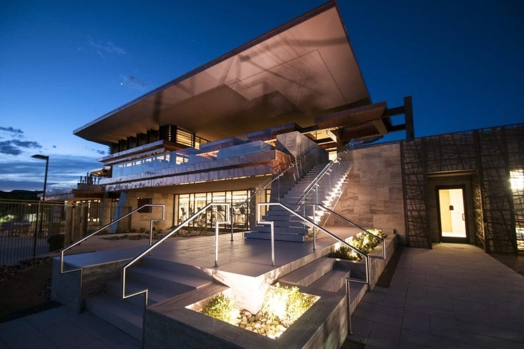 Modern building with large overhanging roof, illuminated steps, and glass walls, photographed at dusk with lights highlighting architectural features.