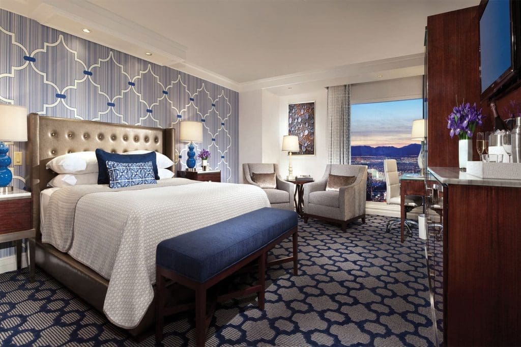 Modern hotel room with a large bed, blue bench, patterned wallpaper and carpet, seating area by a window, and a view of the city skyline at sunset.