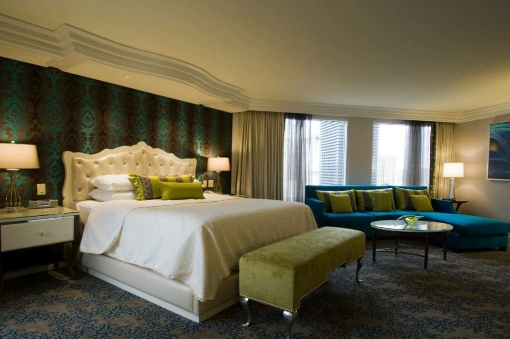 A hotel room with a large bed, decorative pillows, two nightstands with lamps, a bench, a blue sofa, a glass coffee table, and windows with city views.