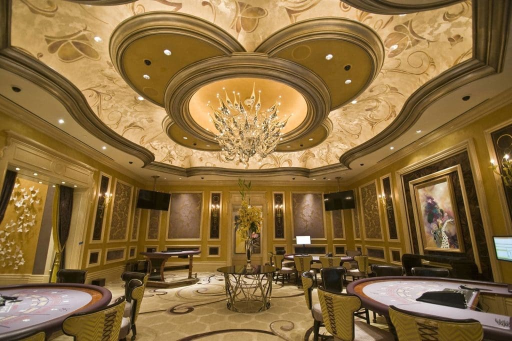 Elegant casino gaming room with ornate ceiling, chandelier, round tables, decorative wall art, and plush chairs arranged around the room.