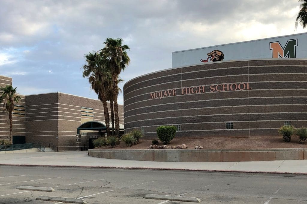 Curved brick building with "Mojave High School" sign, palm trees, and school mascots on the wall, under a cloudy sky.