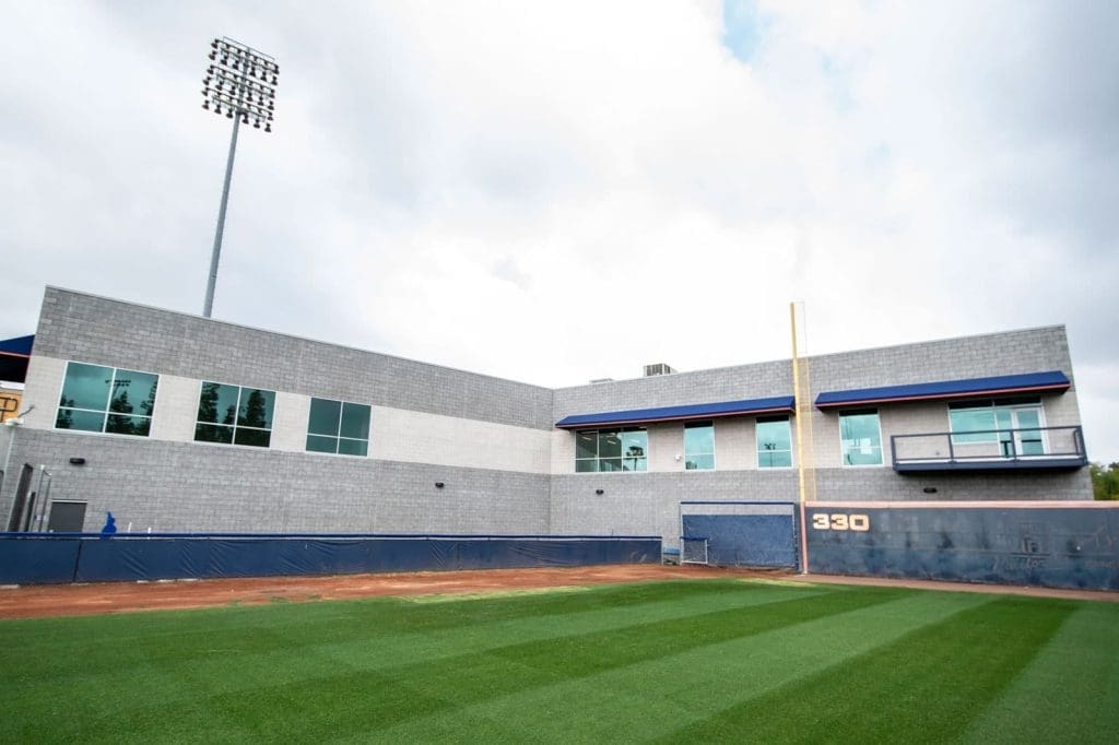 A view of a modern gray building with large windows behind the right outfield wall of a baseball field, featuring a 330-foot distance marker and a tall light pole.