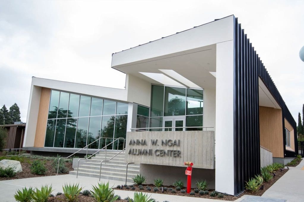 A modern building with large windows and a sign reading "Anna W. Ngai Alumni Center" above the entrance, surrounded by landscaped plants.