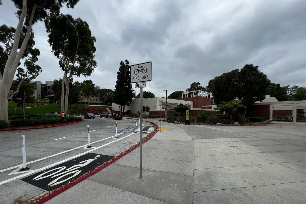 A bike lane with a sign reading "Bike Lane Begin" runs alongside a road bordered by trees, grass, and brick buildings under a cloudy sky.