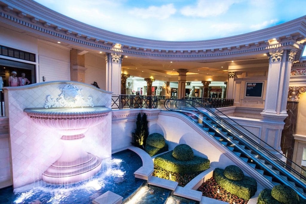 Luxurious indoor space featuring a large decorative fountain, manicured bushes, escalators, and classical architectural details with columns and ornate moldings.