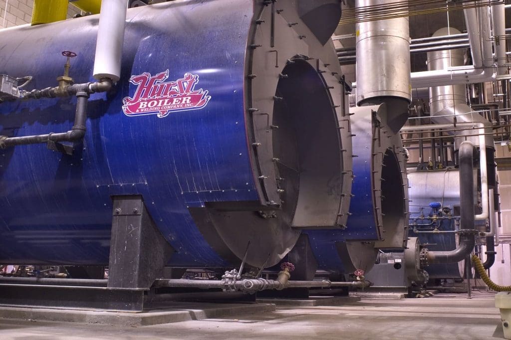 Large industrial boilers with pipes and valves are lined up in a facility. The nearest boiler has a red “Hurst Boiler” logo.