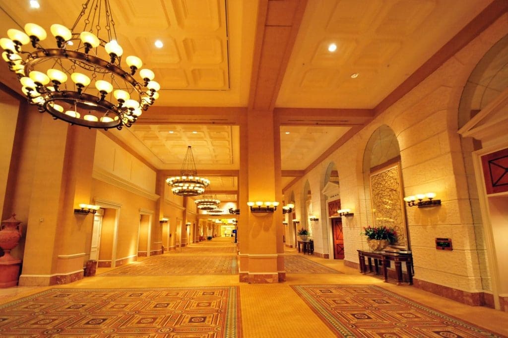 Spacious, well-lit hotel corridor with chandeliers, large columns, patterned carpet, and decorative wall features.