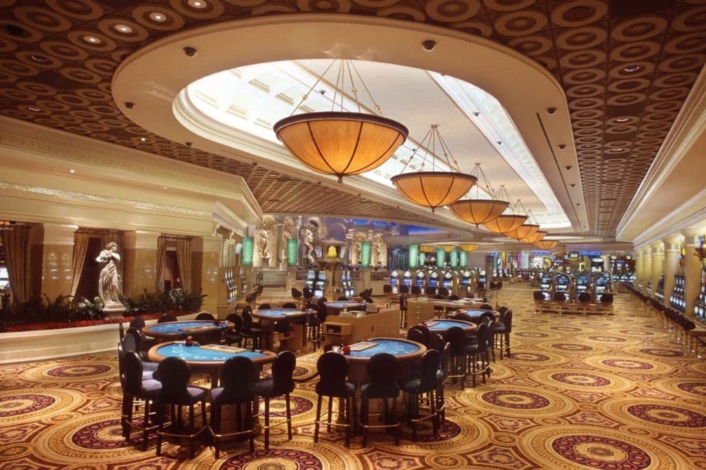 Casino interior with round tables, slot machines, ornate ceiling lights, decorative statues, and a patterned carpet. No people are present.