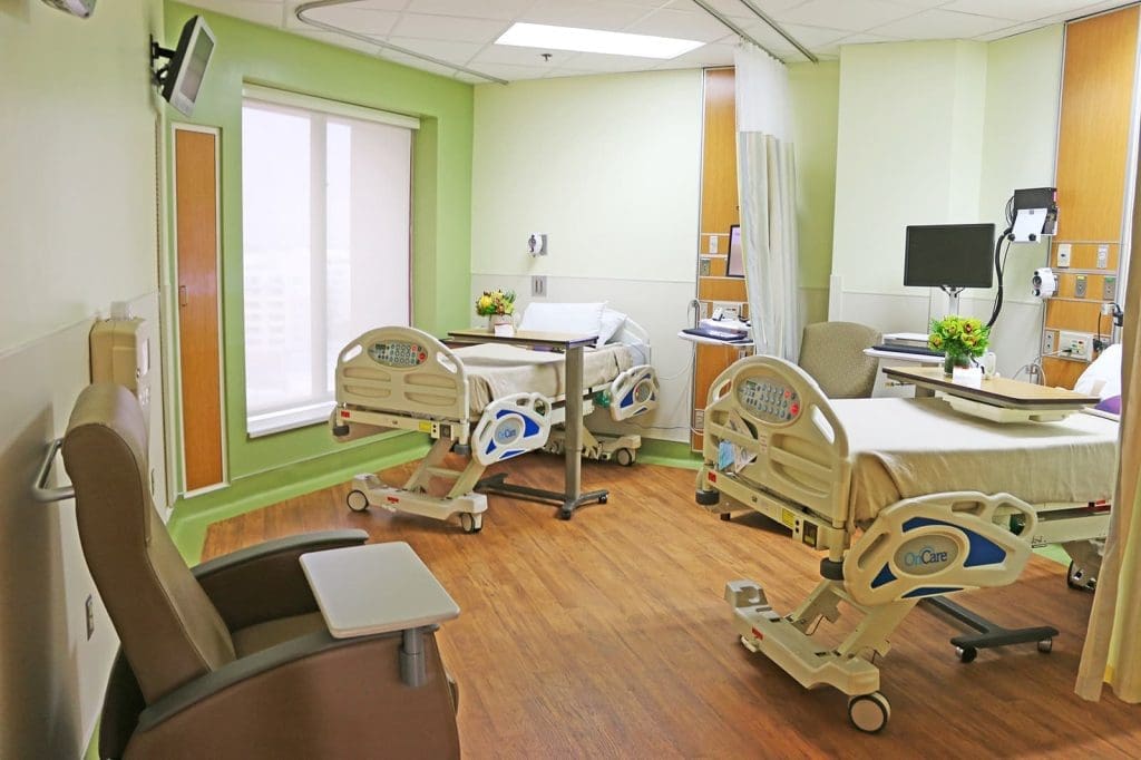 A modern hospital room with two empty adjustable beds, medical equipment, chairs, and flowers on bedside tables, featuring light green walls and wood-style flooring.