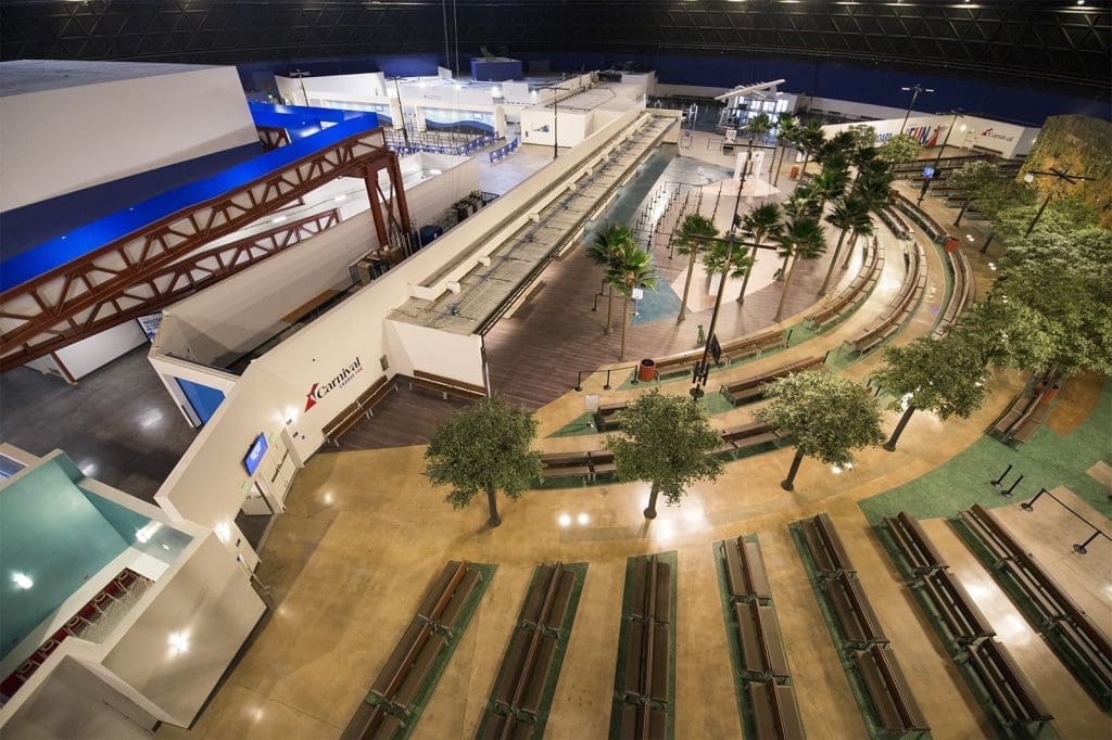 A spacious indoor area with benches, artificial trees, curved walkways, and building facades, resembling an airport or transportation hub.