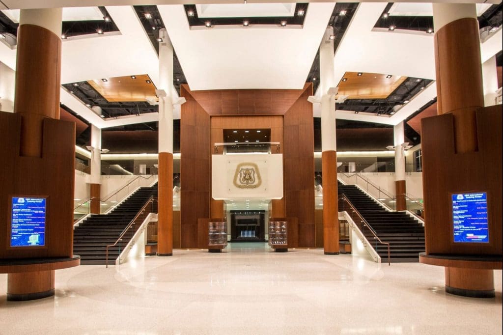 Spacious, modern lobby with high ceilings, wood paneling, two large staircases, digital information screens, and an emblem displayed above the central entrance.