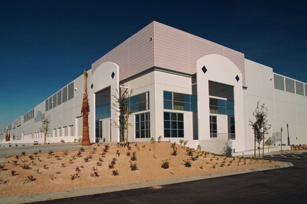 A large, modern industrial building with white exterior walls, large windows, minimal landscaping, and empty parking spaces under a clear sky.