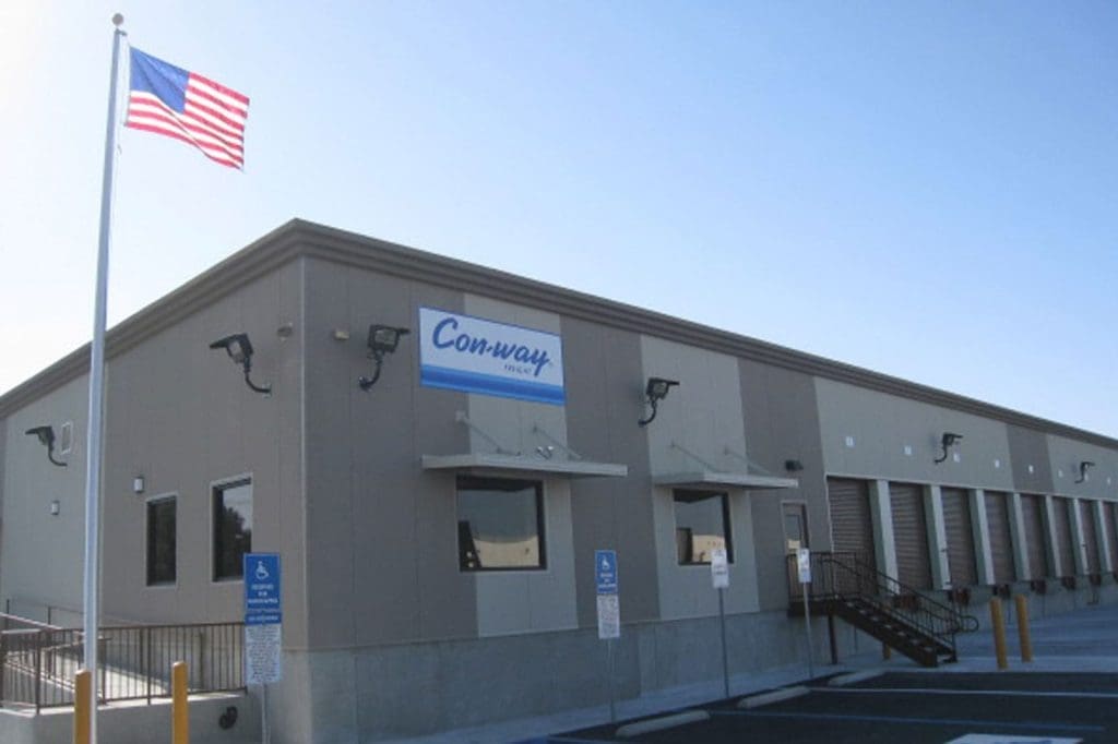 A beige industrial building with a "Con-way Freight" sign, an American flag, multiple loading docks, and designated accessible parking spaces in front.