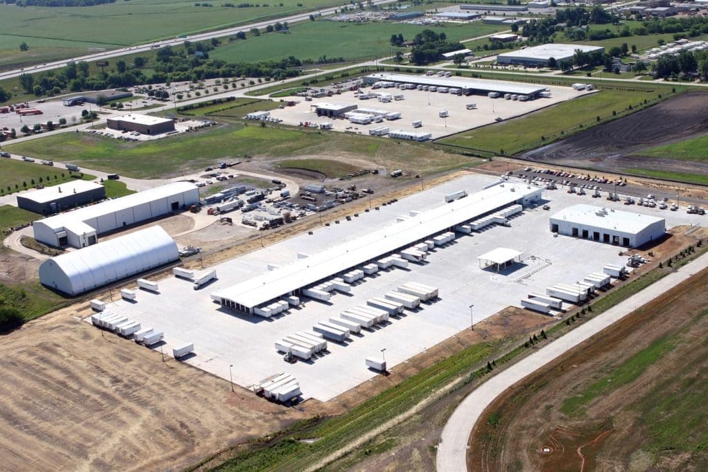 Aerial view of a large industrial facility with multiple warehouses, parked trucks, and open storage areas, surrounded by green fields and nearby buildings.