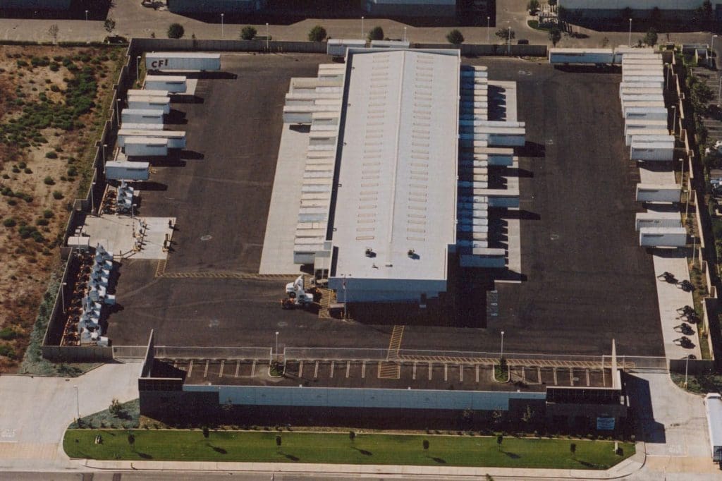 Aerial view of a fenced warehouse facility with multiple truck trailers parked around the building and an adjacent parking lot in front.