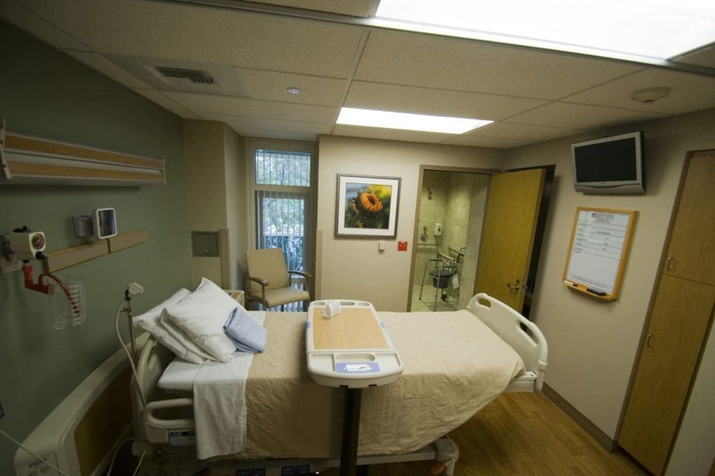 A hospital room with a neatly made bed, medical equipment on the wall, a TV, a whiteboard, a chair by the window, and a framed flower picture on the wall.