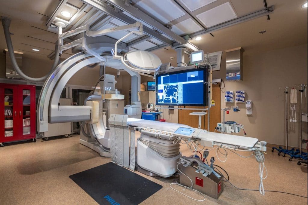 Modern hospital angiography suite with advanced imaging equipment, monitors displaying medical scans, and various medical devices arranged around the patient table.