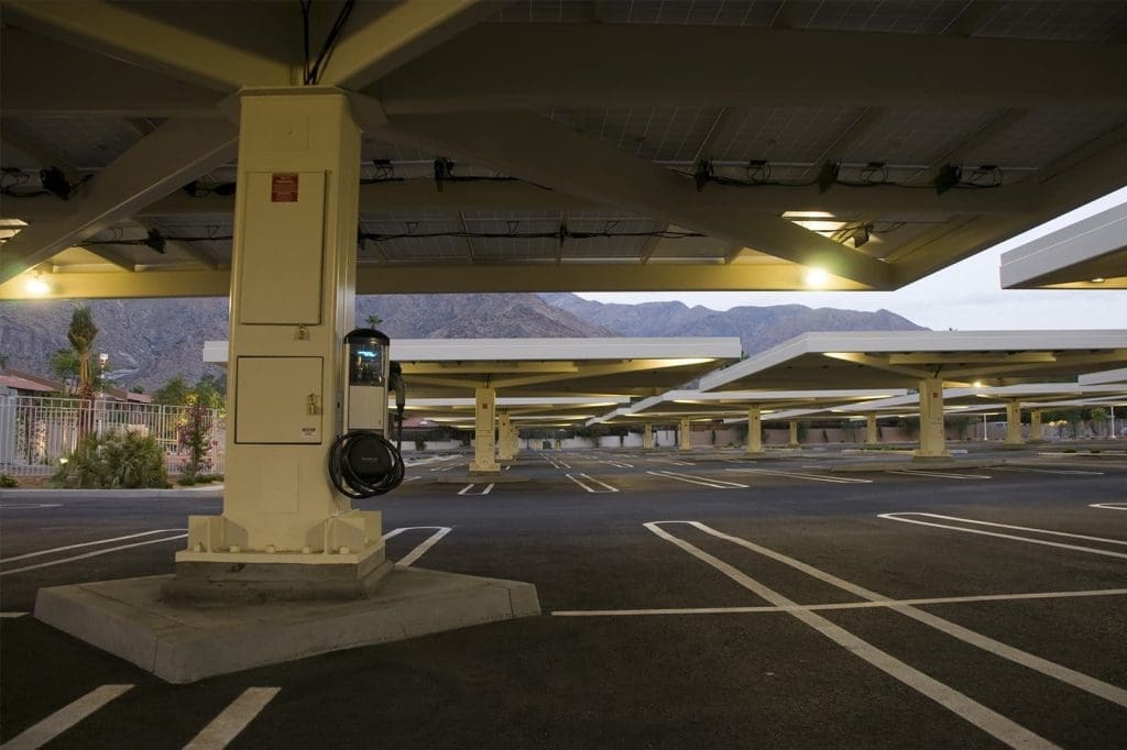 A mostly empty parking lot with solar panel canopies and an electric vehicle charging station attached to a support column. Mountains are visible in the background.