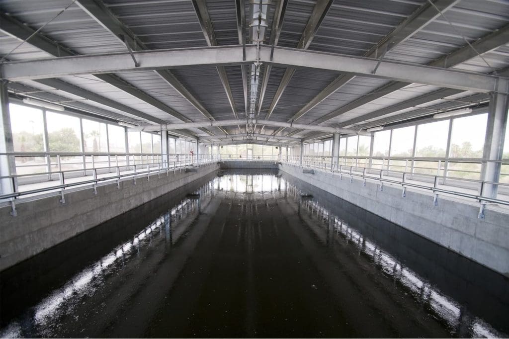 A large, covered water treatment facility with a concrete basin and metal roof, featuring railings along both sides and water pooled at the bottom.