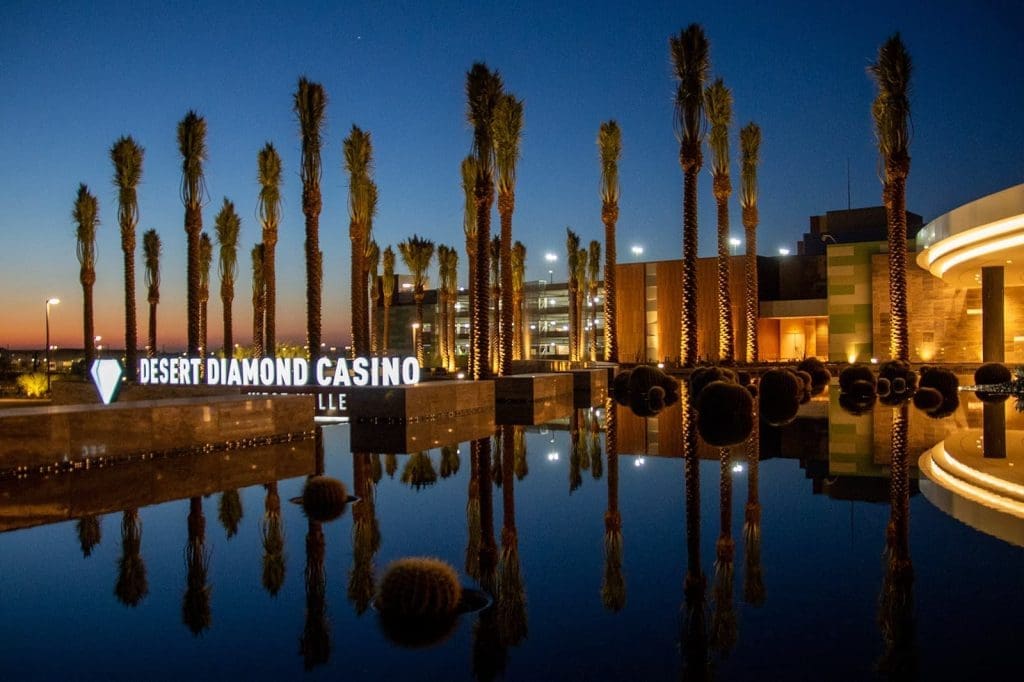 Desert Diamond Casino entrance at dusk with palm trees reflected in a calm water feature and illuminated signage.