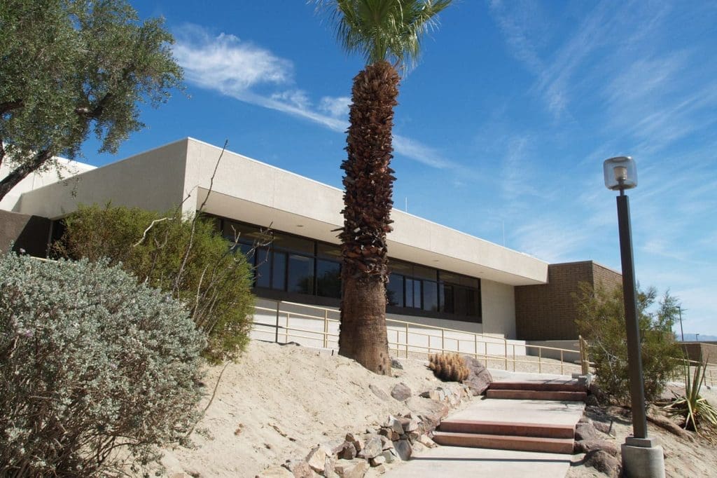 A modern, single-story building with large windows, surrounded by desert plants and a palm tree, under a blue sky with scattered clouds.