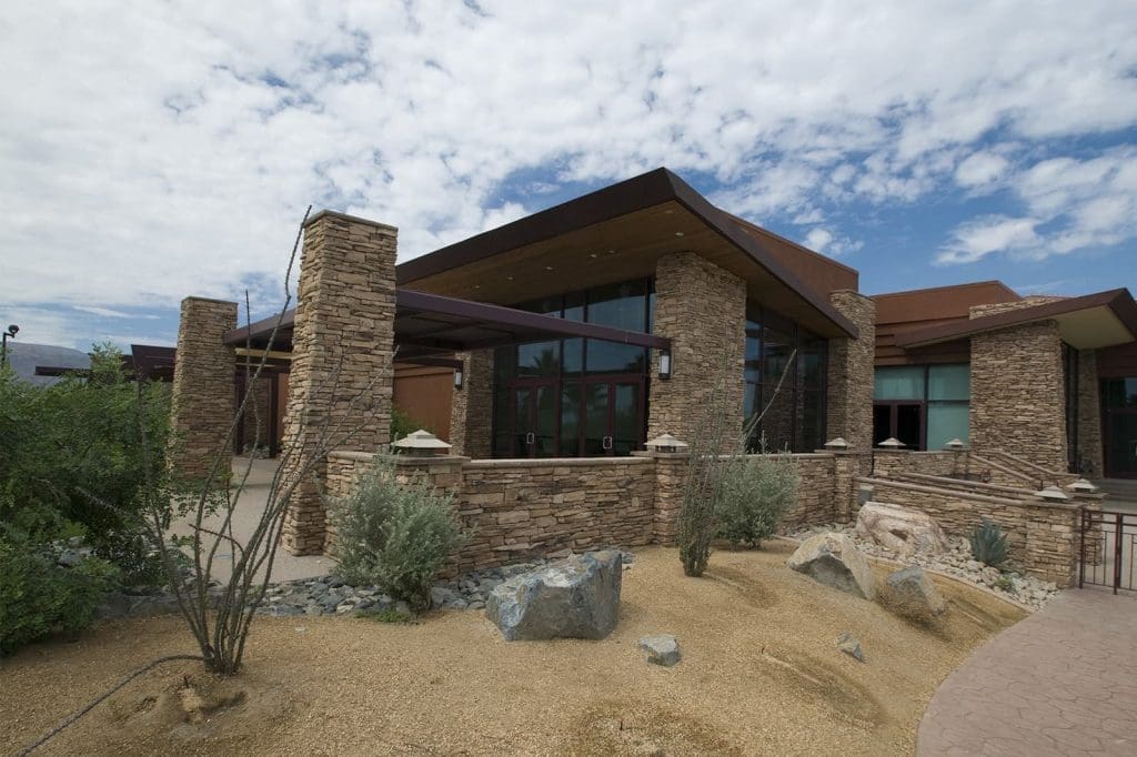 Modern building with stone columns and walls, large glass windows, and a landscaped area featuring desert plants and rocks under a partly cloudy sky.