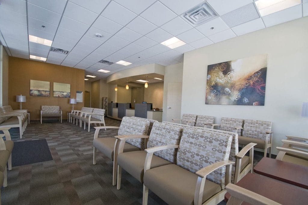 A modern waiting room with patterned chairs, neutral-colored walls, abstract artwork, and a reception desk in the background. The space is empty.