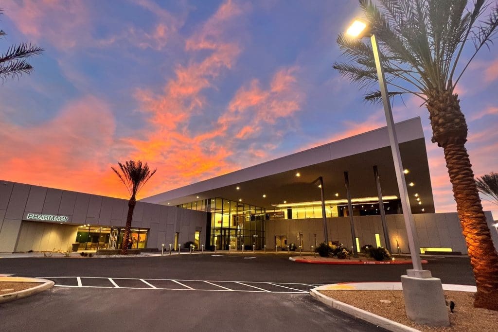 Modern building with large glass windows and a pharmacy sign, illuminated at dusk with palm trees and a vibrant sunset sky in the background.