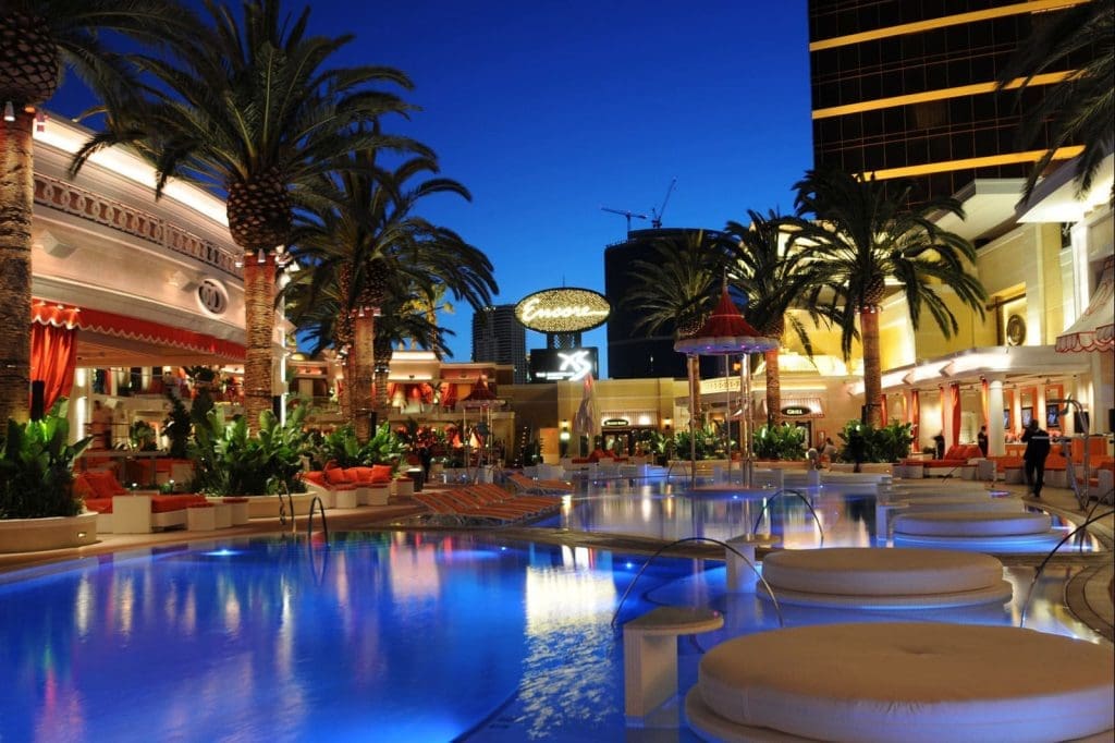 A brightly lit outdoor pool area at night with palm trees, lounge chairs, and the illuminated Encore hotel sign in the background.