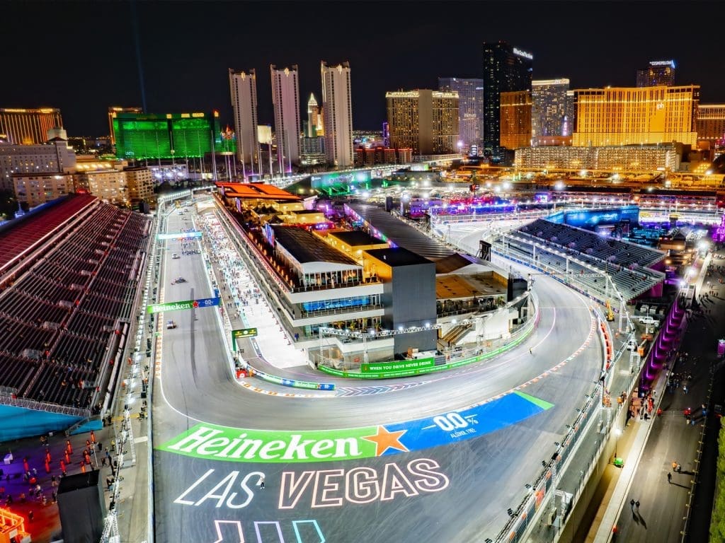 Aerial view of a nighttime Formula 1 racetrack in Las Vegas, surrounded by grandstands and lit-up city buildings, with "Heineken Las Vegas" painted on the track.