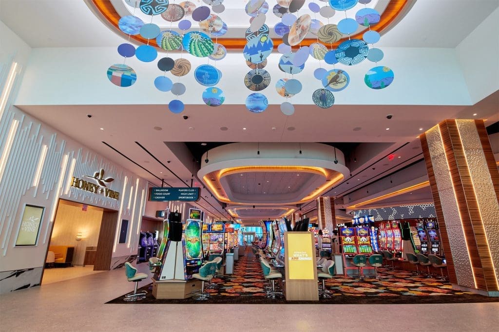 Casino interior with colorful slot machines, modern lighting, suspended circular art pieces on the ceiling, and the entrance to a restaurant called Honey & Vine on the left.