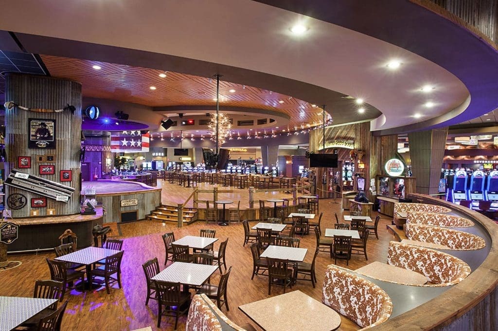 Spacious casino interior with empty dining tables, curved booths, slot machines in the background, wood flooring, and ambient lighting.