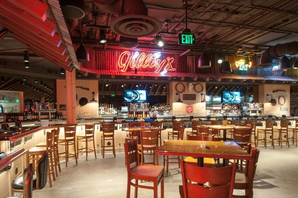 Interior of a spacious bar with wooden tables and chairs, a well-stocked bar, neon signs, and televisions mounted on the back wall.