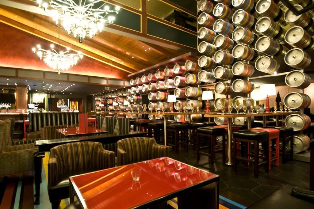 Modern restaurant interior with rows of metal beer kegs on the wall, red tables, striped chairs, bar stools, and decorative chandeliers.