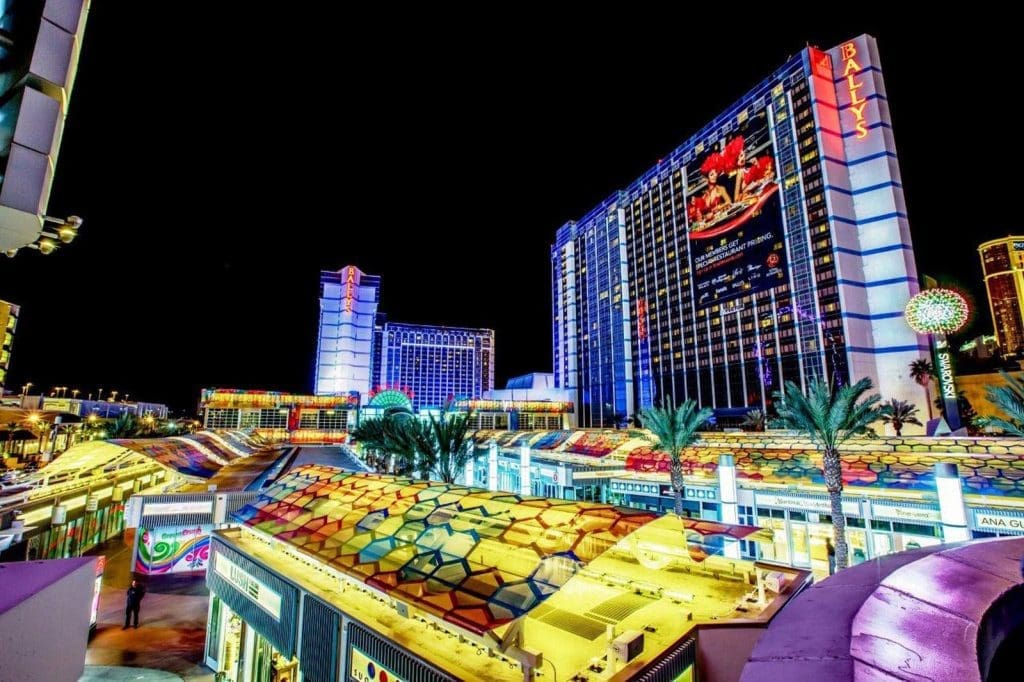Brightly lit hotels and casinos, including Bally's, with neon lights and palm trees along a lively street at night in Las Vegas.