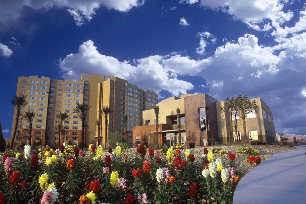 A modern multi-story building stands behind a landscaped garden filled with colorful flowers and palm trees under a partly cloudy sky.