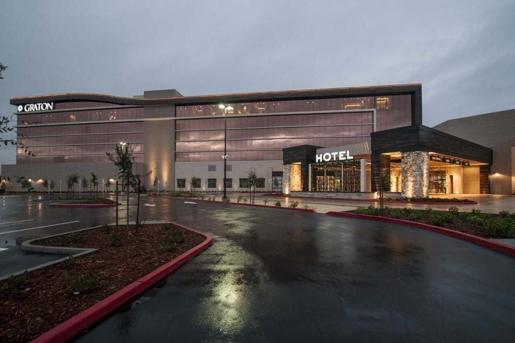 Modern hotel building with large glass windows, labeled "HOTEL" in illuminated letters, and an adjacent structure with the name "GRITON" at dusk. Wet pavement in front.