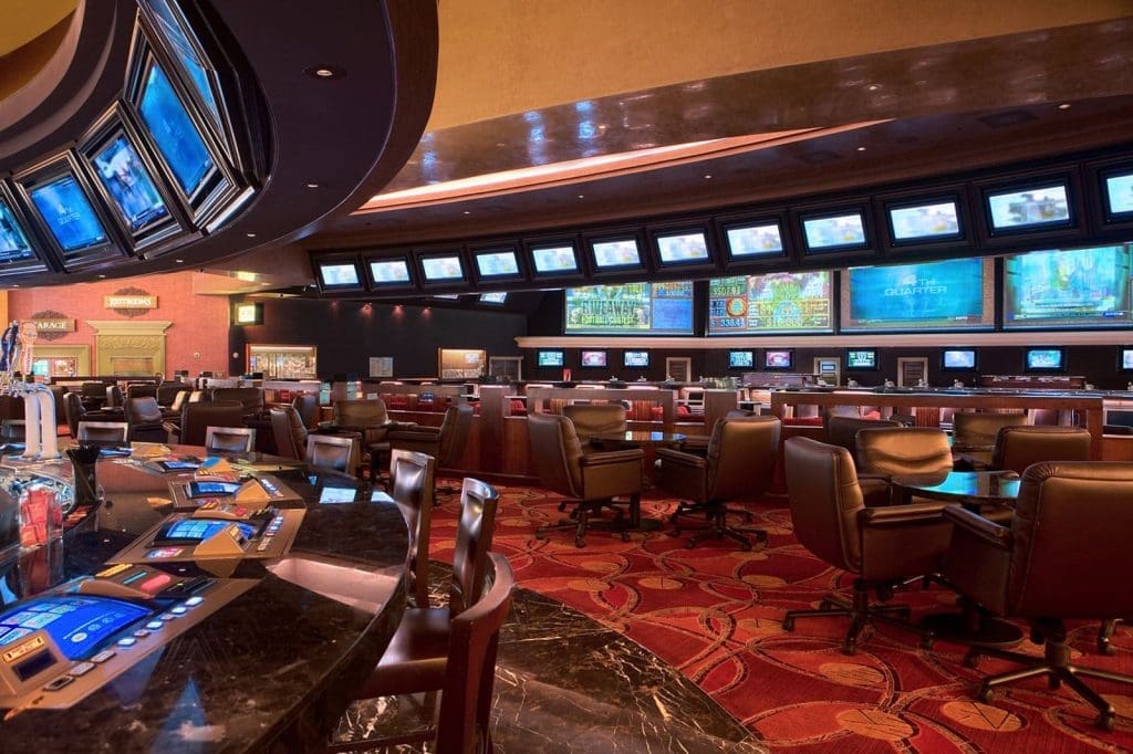 A casino sportsbook area with rows of chairs, electronic betting terminals, and multiple large screens displaying sports events and odds.