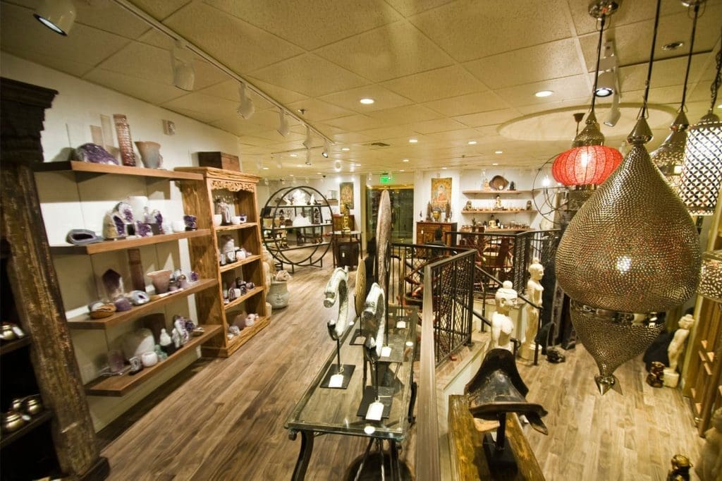 A well-lit store interior displays shelves of crystals, statues, and decorative items on wood flooring, with large ornate lamps hanging from the ceiling.