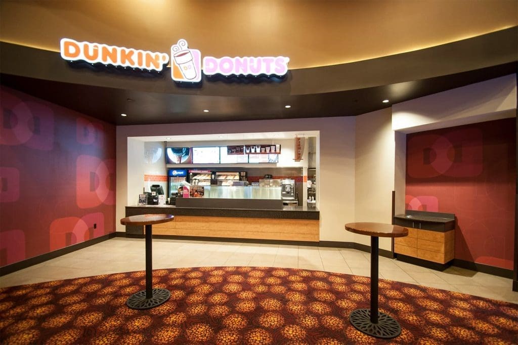 Dunkin' Donuts storefront with counter, menu boards, and coffee machines; two standing tables and patterned carpet in the foreground.