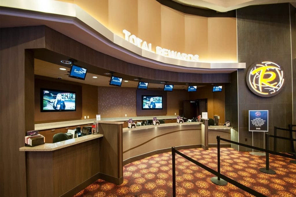 A casino rewards desk with multiple service windows, computer screens, overhead monitors, and a rope barrier on a patterned carpet.