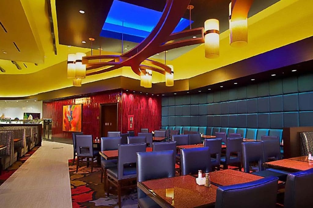Modern restaurant interior with blue padded booths, dark wooden tables, and contemporary lighting fixtures under a yellow ceiling with a blue skylight. No people are present.