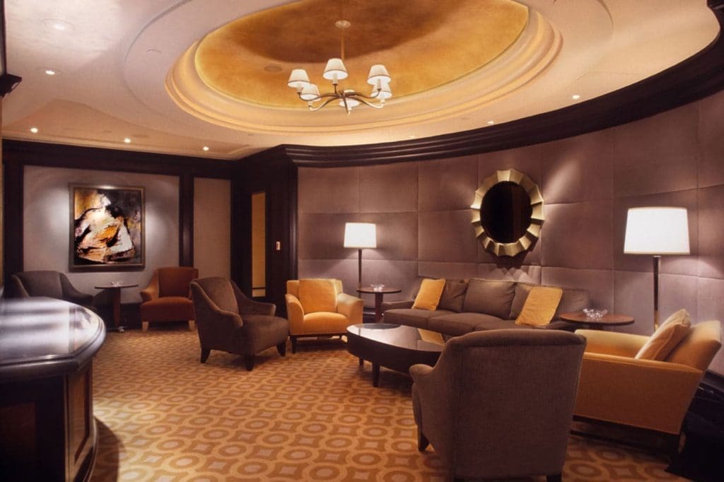 Modern lounge with armchairs, sofas, lamps, a circular mirror, abstract wall art, and a round ceiling detail with a chandelier and yellow lighting.